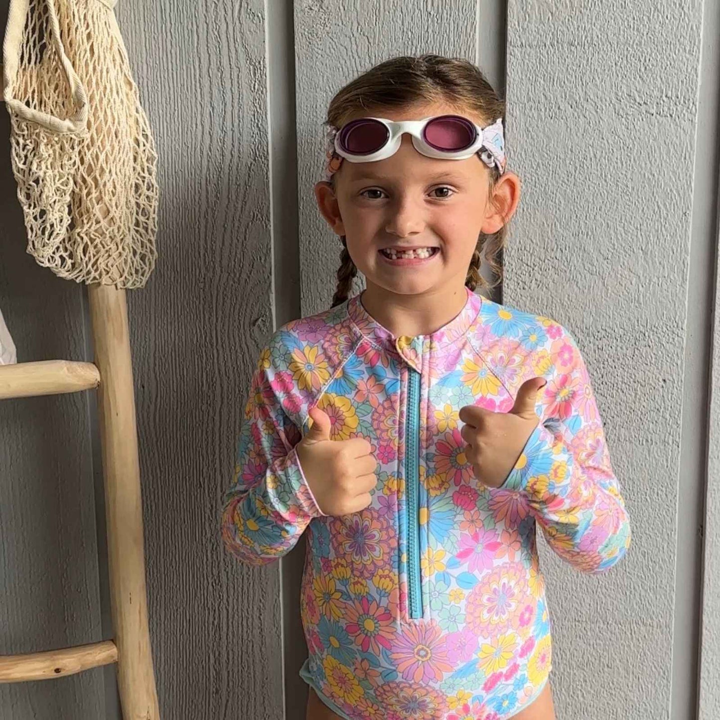 Child in a colorful swimsuit wearing colorful smiley face fabric swim goggles on top of her head and giving two thumbs up - Snappy Goggles