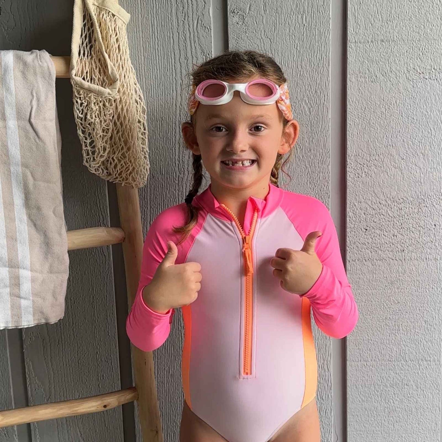 Child wearing a swimsuit with floatable goggles, giving thumbs up in front of a wooden ladder and towel - Snappy Goggles