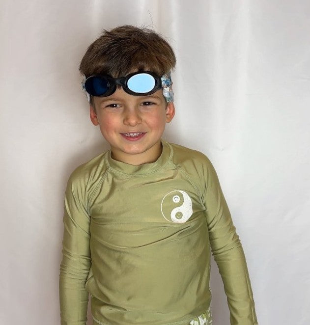Child wearing a green long-sleeve shirt with a yin-yang symbol with fabric swim goggles on top of his head smiling. The gogges don't pull hair and are floatable. - Snappy Goggles