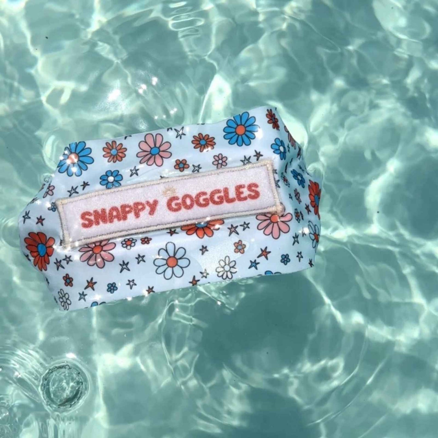Patriotic Floral-patterned fabric strap swim goggles case with 'Snappy Goggles' text floating in clear water.