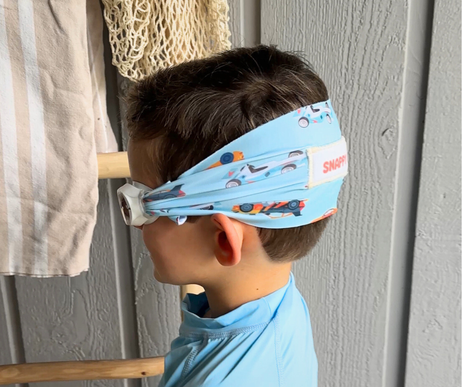Child wearing a blue headband with colorful patterns and text, standing against a gray wall.
