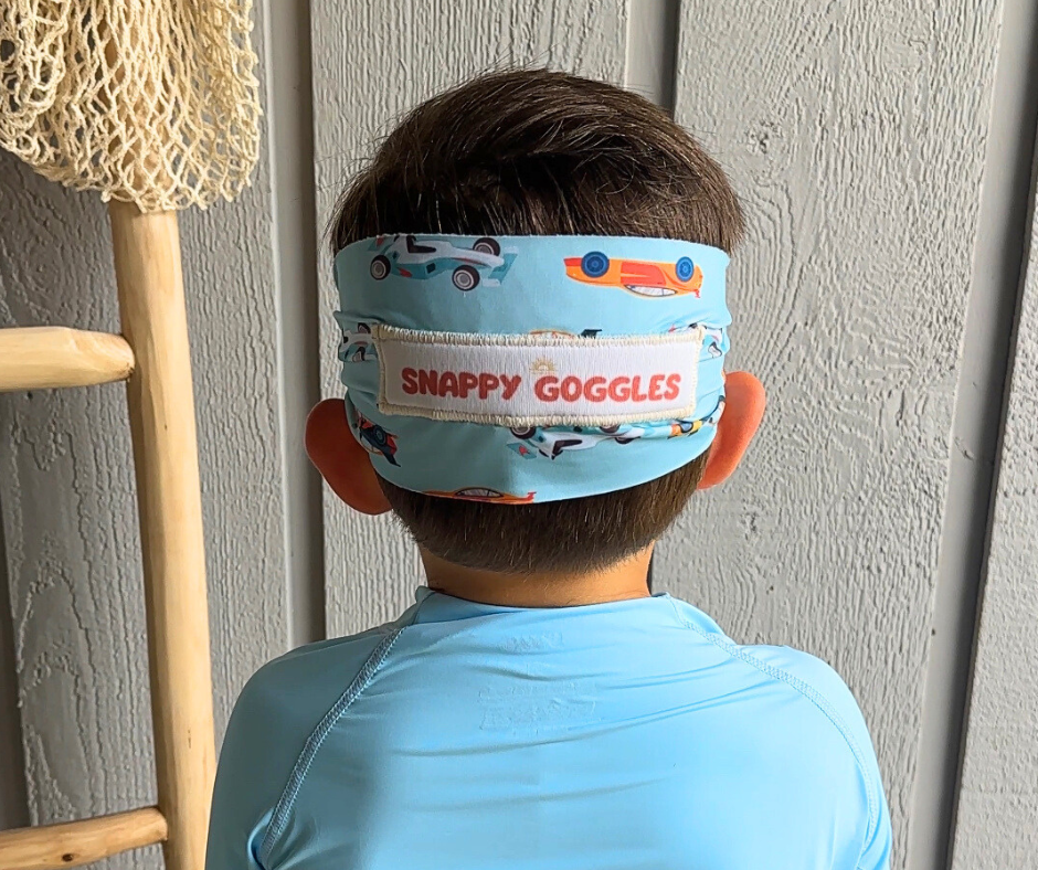 Child wearing a blue 'Snappy Goggles' headband with car designs against a neutral wall.