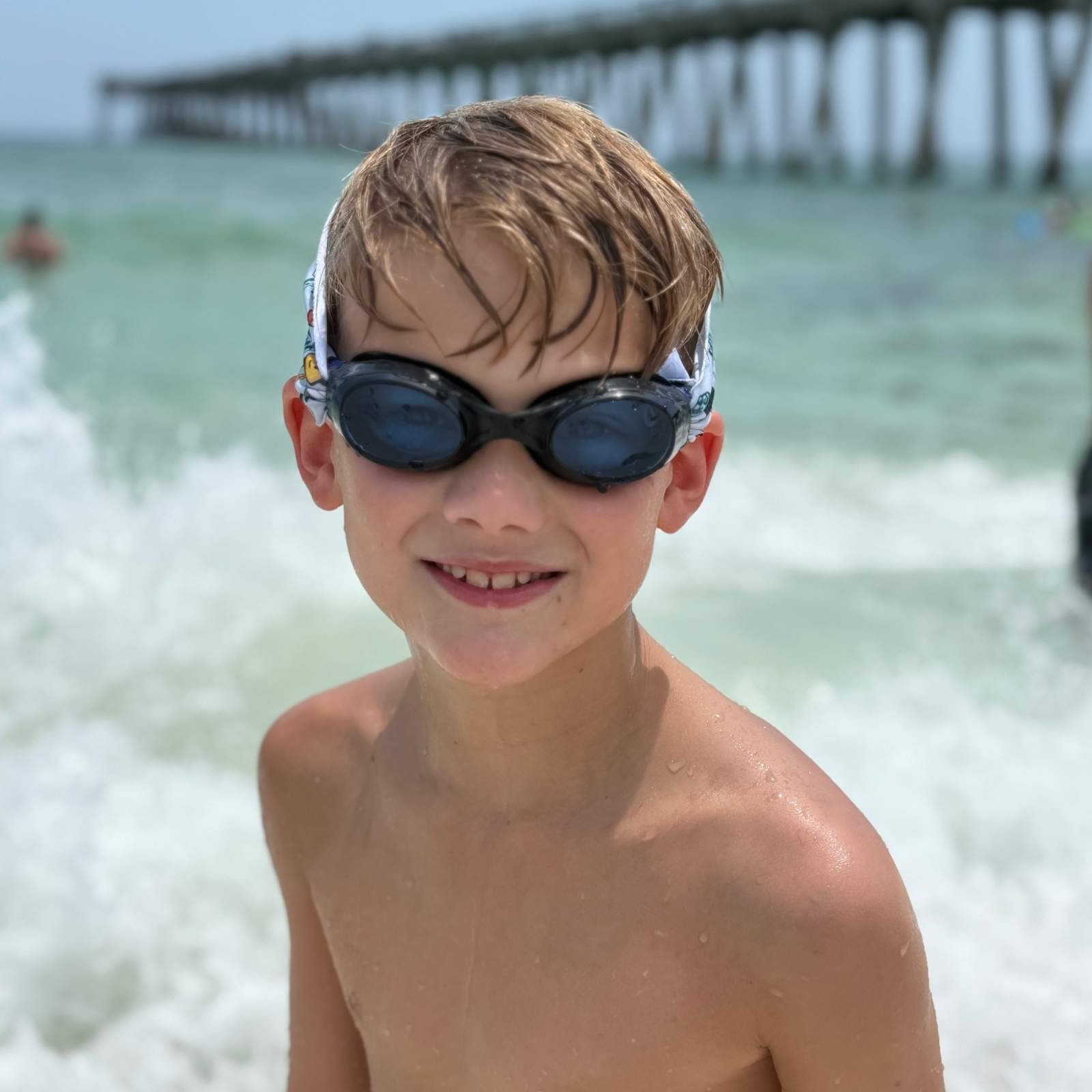Young boy wearing black frame swim goggles with fabric strap at the beach with a pier in the background - Snappy Goggles