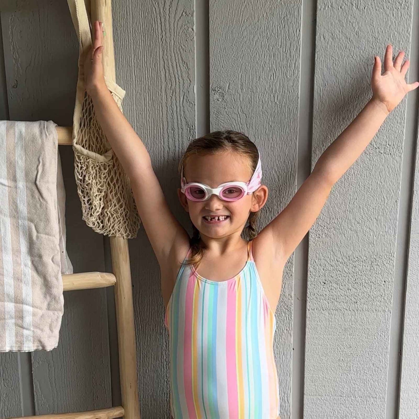 Child in a striped swimsuit standing wearing strawberry fabric swim goggles that don't pull hair -Snappy Goggles