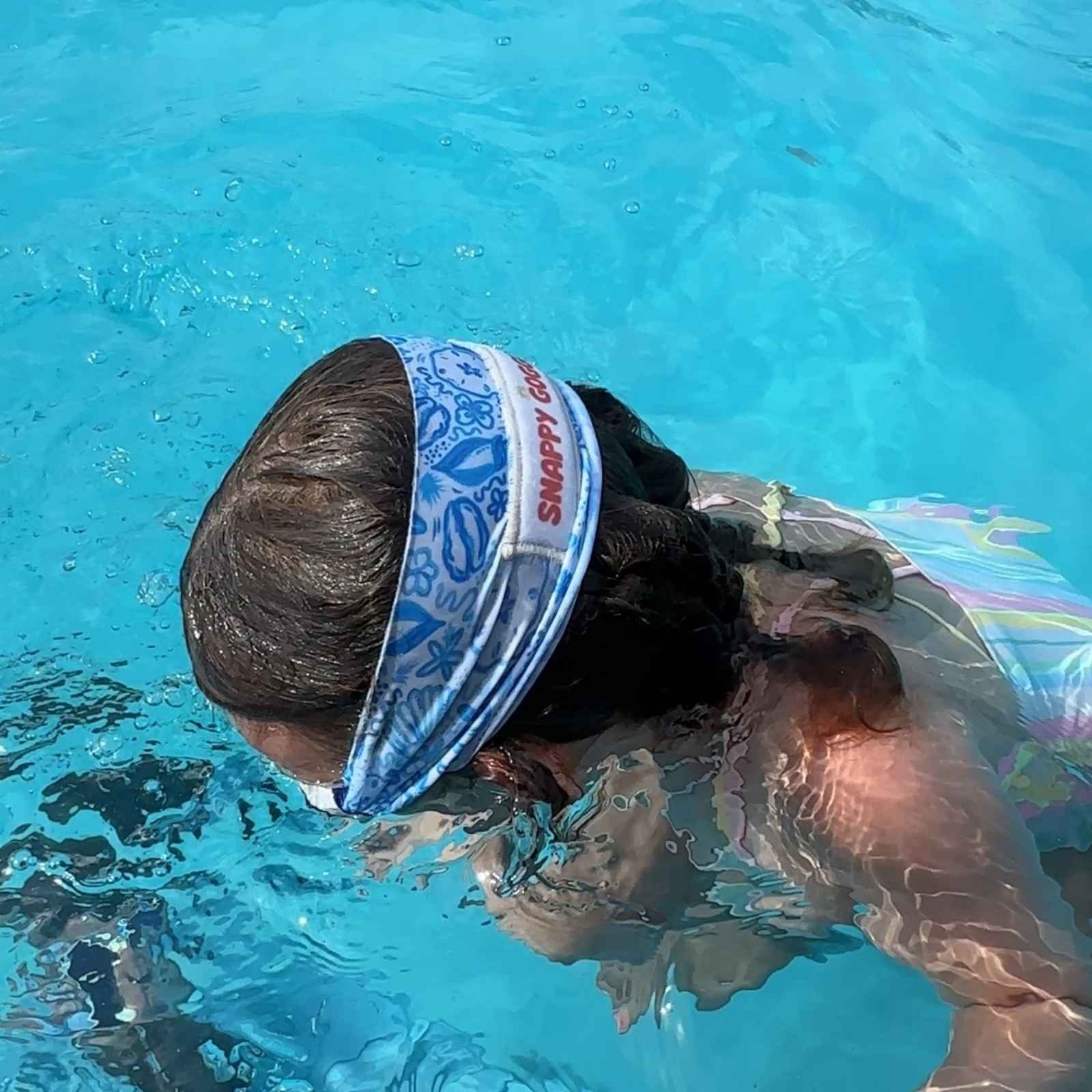 Girl swimming in a pool with a colorful swimsuit and soft, blue seashell fabric strap swim goggles that float - Snappy Goggles