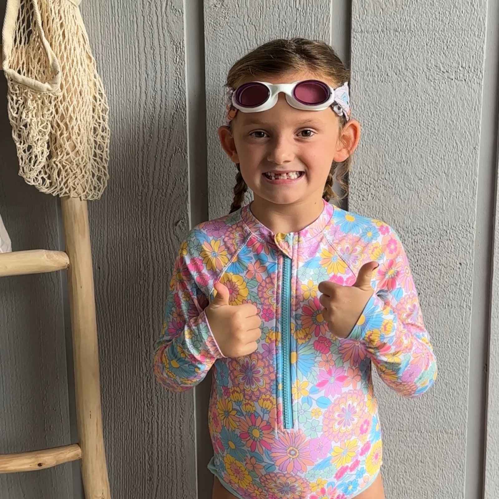 Child in a colorful swimsuit wearing colorful smiley face fabric swim goggles on top of her head and giving two thumbs up  - Snappy Goggles
