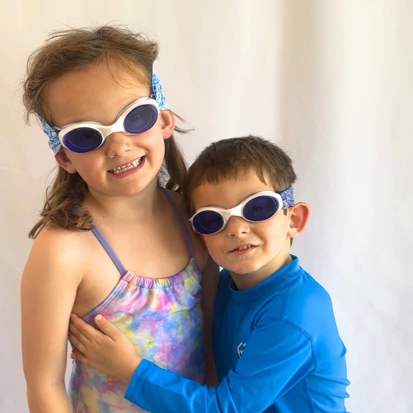Two children wearing fabric swim goggles featuring blue seashells and blue waves - the goggles don't pull hair and are floatable - Snappy Goggles